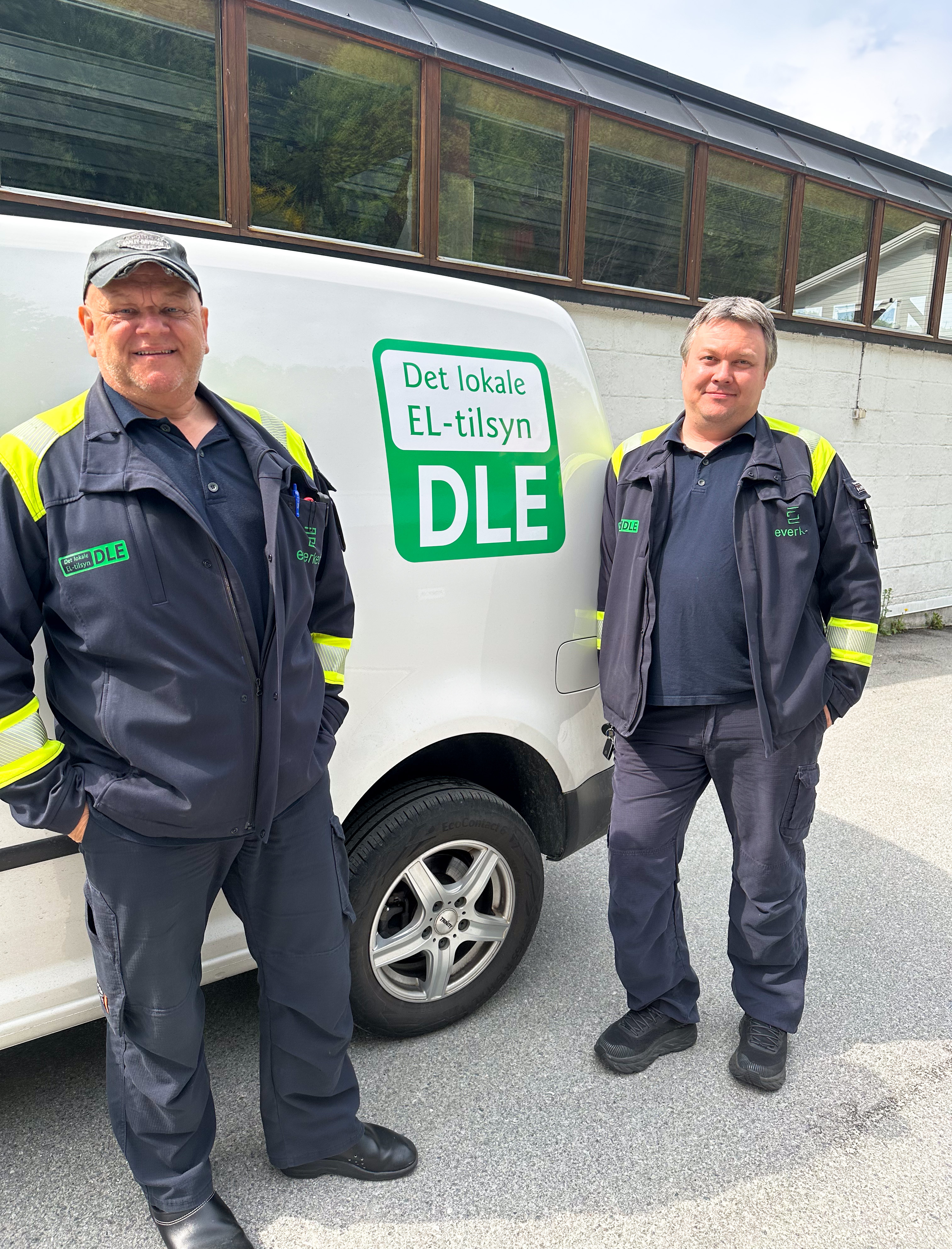 To ansatte i arbeidsklær står ved siden av en hvit firmabil med logoen 'Det lokale el-tilsyn (DLE)'. De er utendørs foran en bygning. Foto.