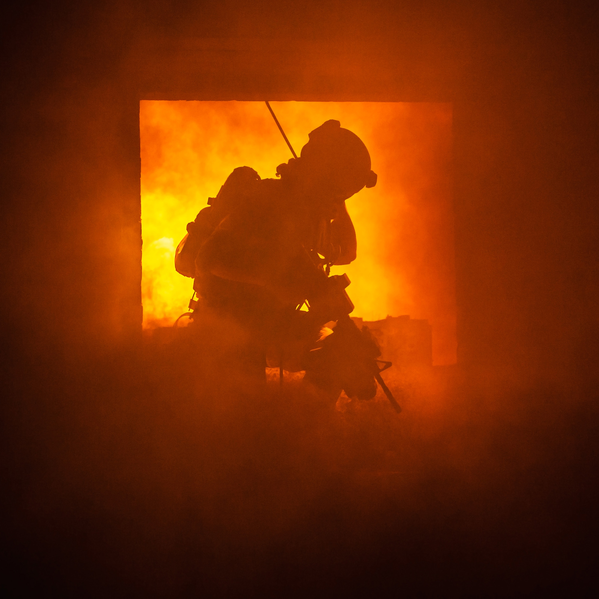 En brannmann i fullt verneutstyr beveger seg gjennom et rom fylt med røyk, med kraftige flammer i bakgrunnen. Personen er delvis silhouettert mot det sterke lyset fra brannen. Foto.
